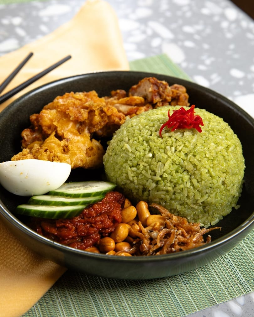 A black bowl of green rice, fried chicken, cucumber, egg, sambal, peanuts, and anchovies.