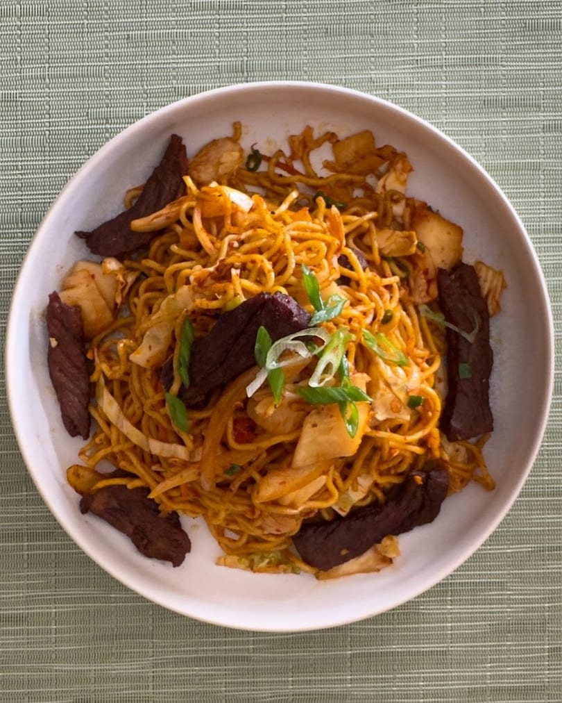 A white bowl filled with noodles, beef strips, cabbage, and green onions on a green placemat.