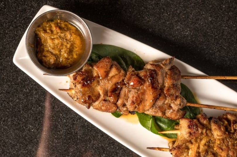 Grilled chicken skewers with a side of peanut sauce on a white rectangular plate with spinach.