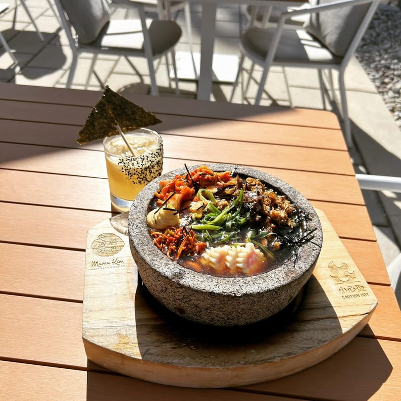 A stone bowl of Mama Kim's ramen and a cocktail with a seaweed garnish on an outdoor table.